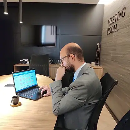 Bruno de Bruno lendo no notebook, com a mão no rosto, na sala de reuniões